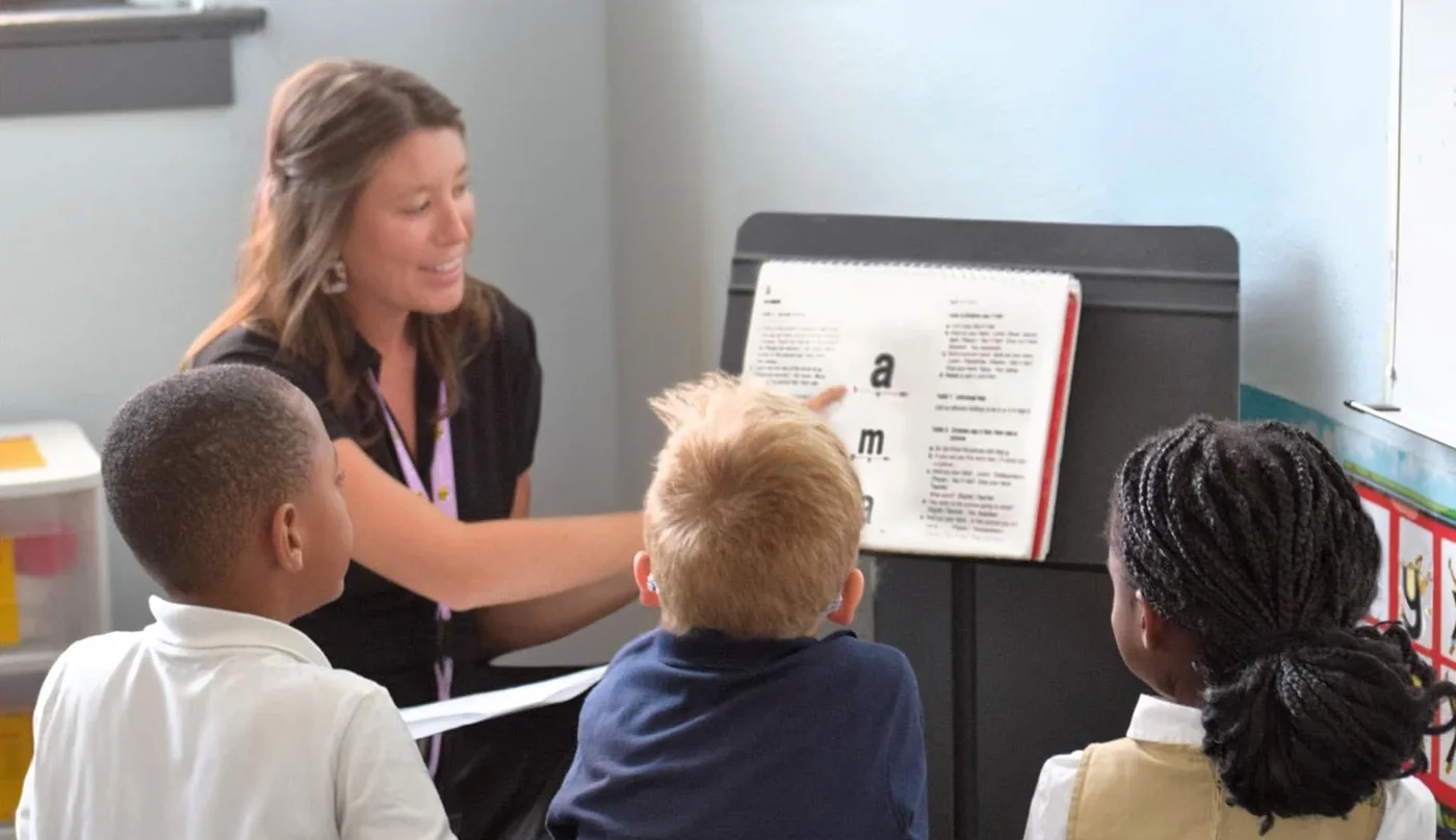 A smiling teacher guides a small group of children through a reading lesson.