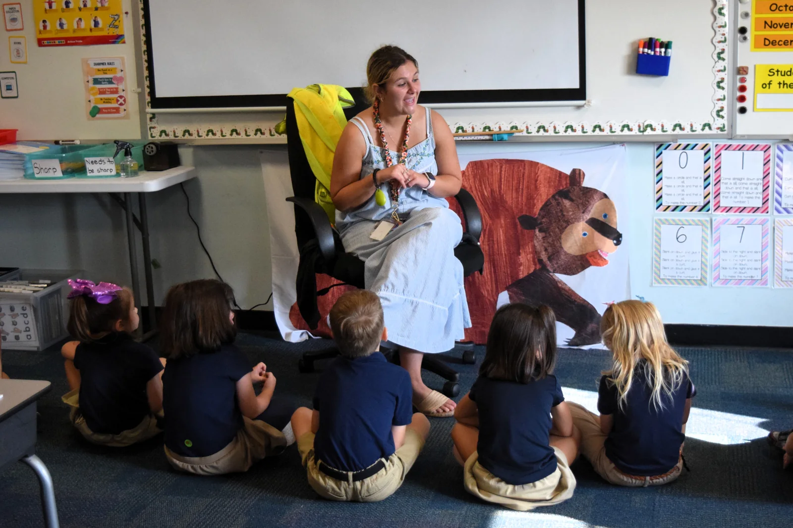 Teacher smiles while leading a classroom activity with students seated in a circle.