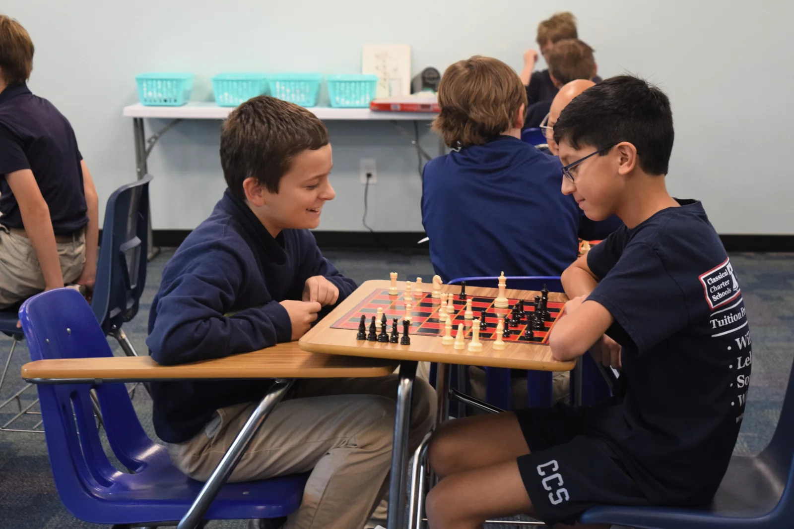 Two students smile while playing chess together in a classroom.